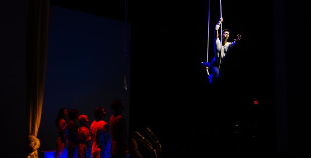 A performer swings from a rope, while other performers stand below on a stage in red light.