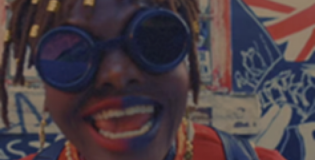 A Black woman with braids, and wearing goggles and red and blue lipstick on the left and right halves of her mouth, smiles wider.