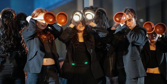 Three people dressed in black hold toilet plungers in front of their faces.