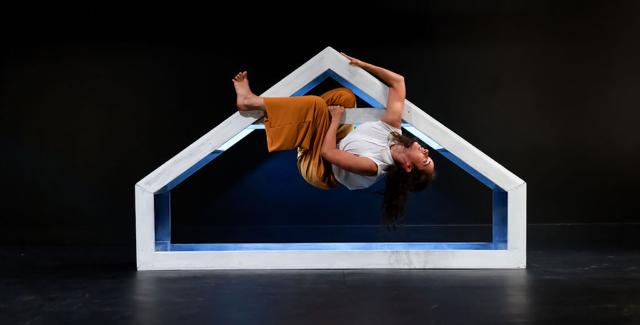 On a stage, a woman hangs from a frame that is shaped like a short house.