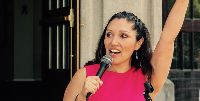 Jennifer holds a mic in one hand and raises her other fist before a crowd of people.