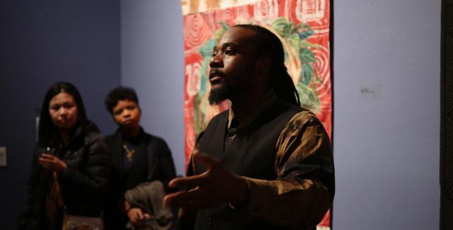 Stephen speaks to a room of people in front of a tapestry.