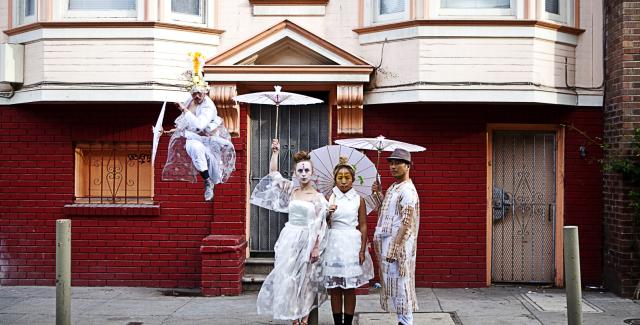 Outside a San Franciscan home, a person dances on a stoop while the others wear white lace and hold white umbrellas.