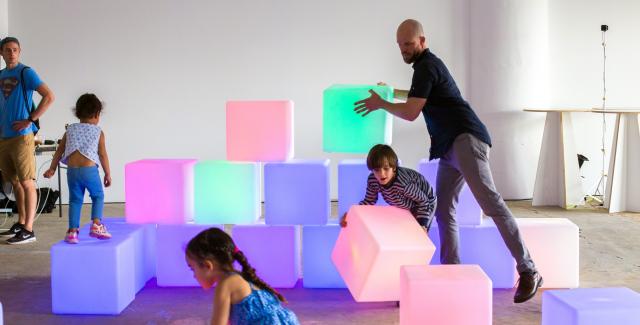 Ryan places a light up boxe about twice the size of a shoeboxe on top of about twenty others with some children.