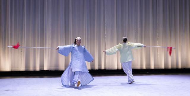 On a stage with a white curtain behind them, two performers hold white sticks with red strings at the end and face each other while walking in opposite directions, but possibly in a circle.