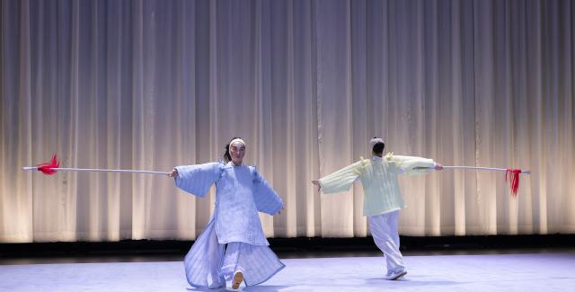 On a stage with a white curtain behind them, two performers hold white sticks with red strings at the end and face each other while walking in opposite directions, but possibly in a circle.