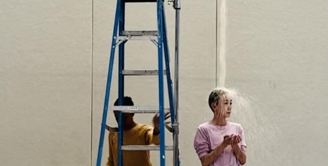 A woman stands behind a ladder while another woman has water dropped on her from an iv bag.