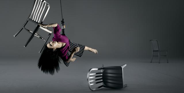 In a gray space, a performer glides while hanging next to a chair that is also strung from the ceiling. other chairs are turned over.