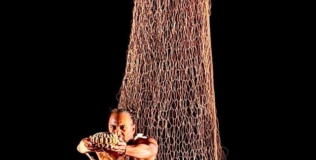 On a dark stage, a person stands in front of a net that drapes from the ceiling.