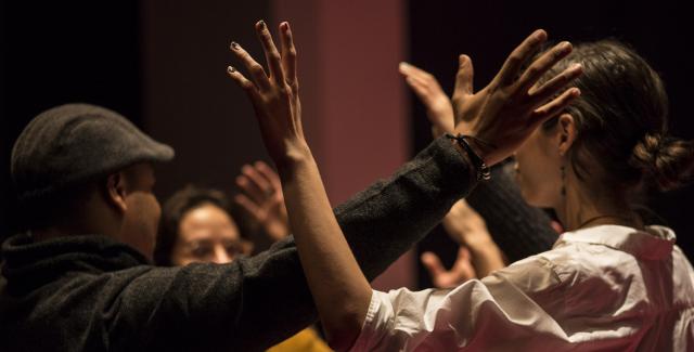 Two people are part of a larger crowd of people with their arms held up and touching each other at the elbows. The crowd is doing this in a circle.