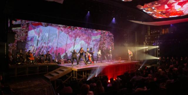 A dark theater with 8 actors facing the audience and a band in the background. Multicolored fabric and projections adorn the backdrop and the ceiling.