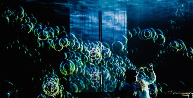 On a stage, a person blows lots of bubbles. Underlit, the bubbles create an oceanic print on the ceiling.