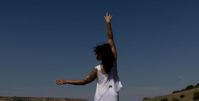 In the desert, a woman. with shoulder-length dark hair and in a white outfit, holds her right hand to the sky and left to the left.