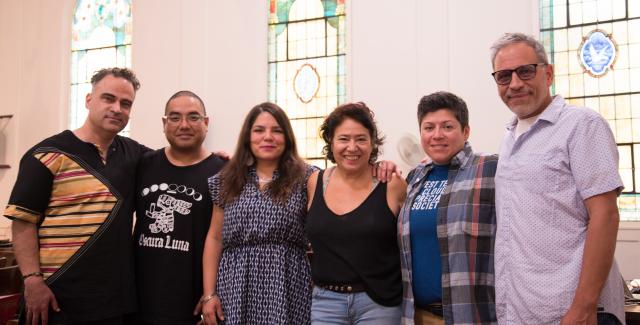 Six people in casual clothing standing arm in arm facing the camera. Large stained glass windows are in the background.