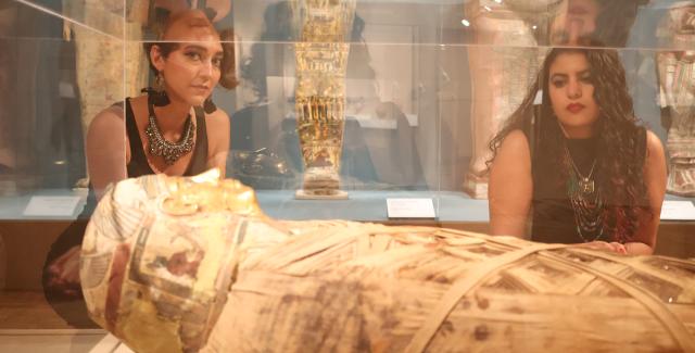 In a museum's glass case, a mummy. Outside the glass, two women peak inside.
