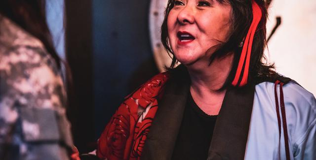 An elder Japanese woman speaks to another person. She stands in front of a gong and has a red ribbon tied to her hair.