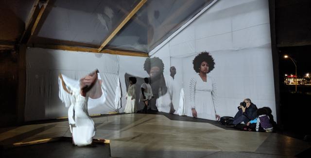 In an outdoor space with white walls and slanted ceiling windows, a dancer holds fabric over herself. A portrait of Black women is projected over her and the white walls.