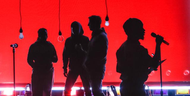 On a stage, with a red backdrop and lightbulbs hanging from the ceiling, four performer silhouettes sing into microphones.