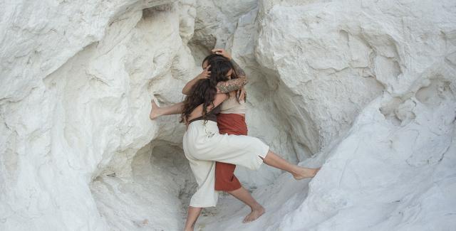 In a white rock formation, two women wrap their bodies around each other. They have brown hair and wear casual attire.