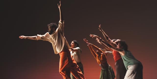 A male dancer leaps up while female dancers behind him reach towards him.