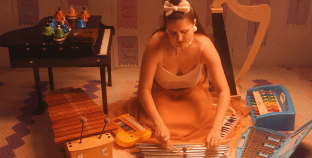 alexa dexa, a queer, non-binary, Sick, Mad, and Disabled white person with light olive skin tones and long golden brown hair, wears a silk gown with a white top and flowing peach skirt and kneels on the floor playing their toy chimes. Surrounding them from left to right are members of their beloved toychestra: a 37-key Schoenhut baby grand toy piano, 20 chromatic desk bells, kalimba, alto xylophone, elephant toy piano, vintage noisemaker, toy glockenspiel, melodica, tap-a-tune xylo-keyboard, and toy harp.