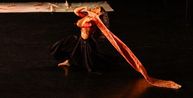 A performer in a dark skirt drags a piece of orange cloth across a stage.