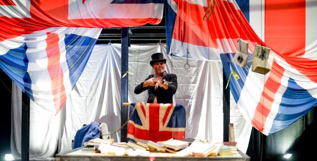 Nora, in a top hat, performs on a stage with British flags.