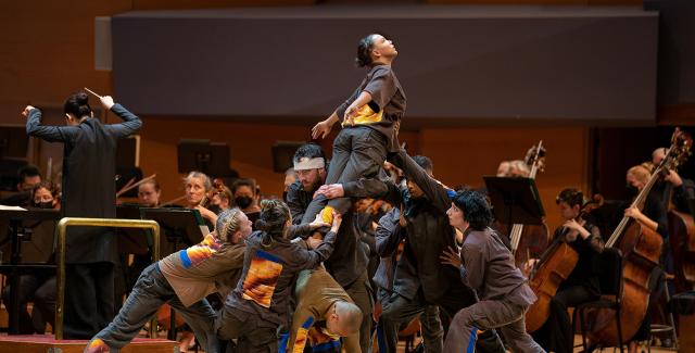 Six dancers hold a woman, who extends her arms, up above them.