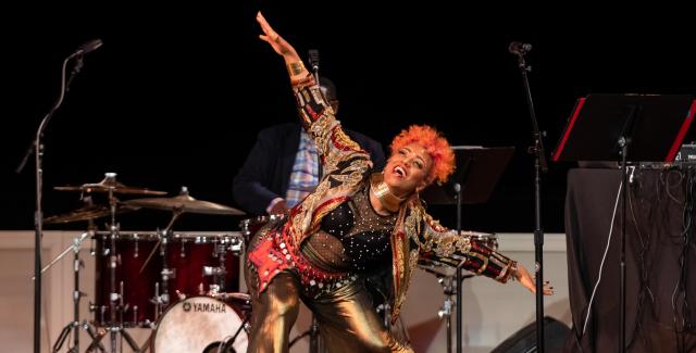 On a stage, between mic stands, a Black woman, with orange hair and gold clothes, bends and holds her arms out wide.