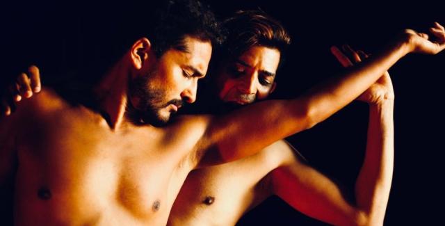 Two shirtless Indian men caress each other in front of a black backdrop.