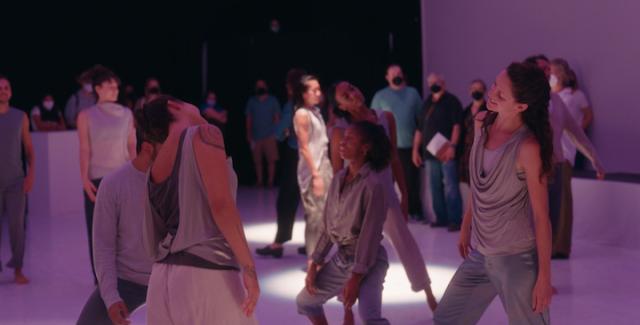 In a gallery space, dancers, wearing creams and grays, slouch. A masked crowd watches.
