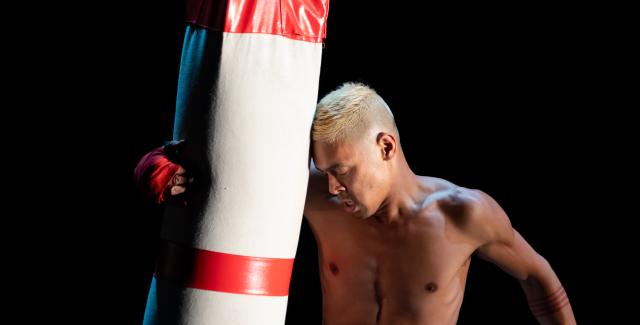 A brown man, with blonde hair, holds a punching bag against his head.