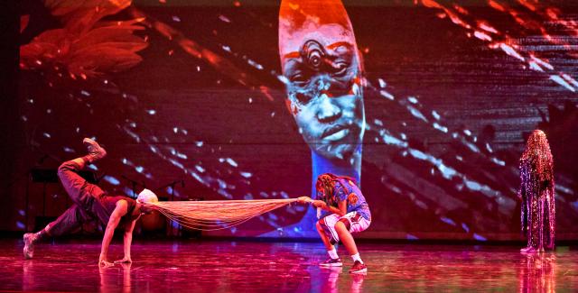 On a stage, a dancer pulls another dancer by their fringe mask, while another looks on. Behind them, a projection of an Afro-Futuristic image, where a person with a long neck and face has three eyes.