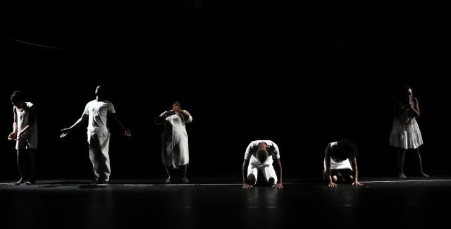 In front of a black backdrop, dancers in white stand in a line in various poses.