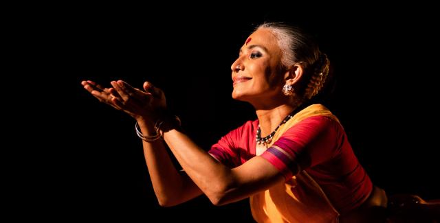 An Indian woman holds her hands up to a spotlight. She wears a sari. 