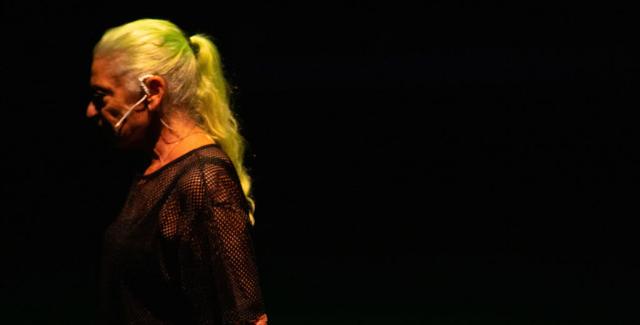 In a spotlight, a feminine person with long blond or yellow hair wears an earpiece and black mesh.