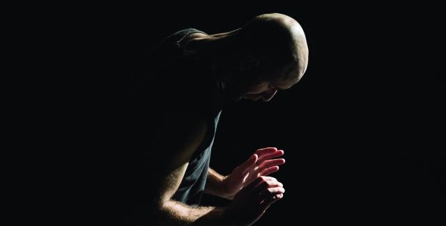 a bald man crouches and lifts his hands up facing out by his chest.