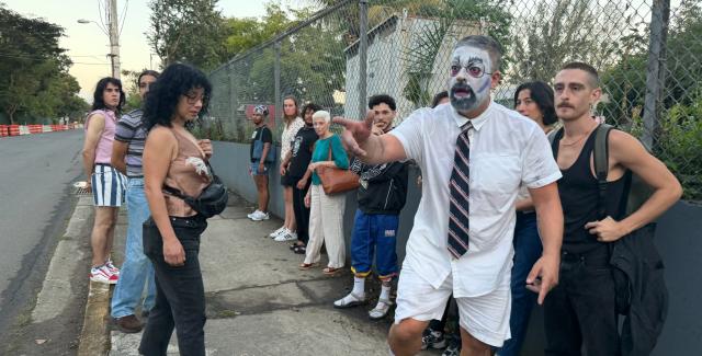 A person whose face has been painted white stands on a street, pointing at something offscreen. Eleven people look in the direction he's pointing.