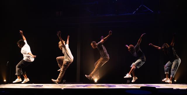 Five performers on a stage are mid leap with their arms raised over their heads and extended outwards.