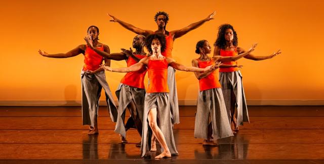 On a stage, with an orange backdrop, six dancers, in orange tank tops and grey skirts, spread their arms.