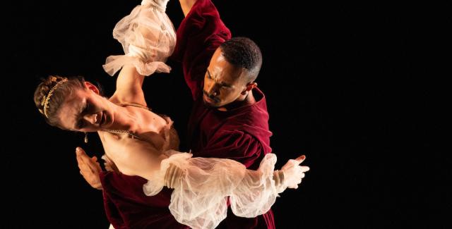 Two people in anachronistic clothing do a waltz in front of a black backdrop.