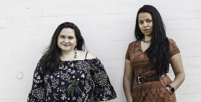 Two women of color pose next to each other on some outdoor steps, in front of white bricks. They both wear heels - one wears a brown wrap dress and brown, heeled boots, while the other wears a flowy black and white top off one shoulder and black denim with grey gladiator sandals.