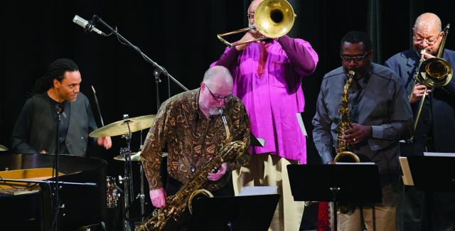 Five men are on stage. Four play brass and one plays drums.