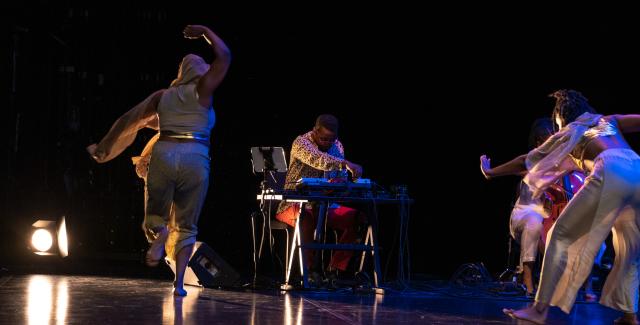 On a stage, Black women dance while a Black man plays the keyboard. Spotlights are also on the stage.