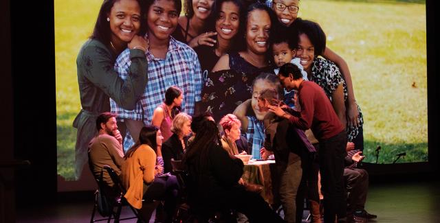 On a stage, six white folks sit around a table. A white man leans over them and gestures with his hands. Behind them is a portrait of a group of people of color.