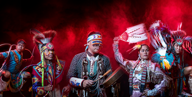 In front of red smoke, five performers in traditional native garb dance and play music.