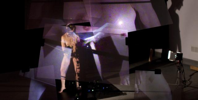 Projections of different parts of a female surround a nude male figure in a dark room.