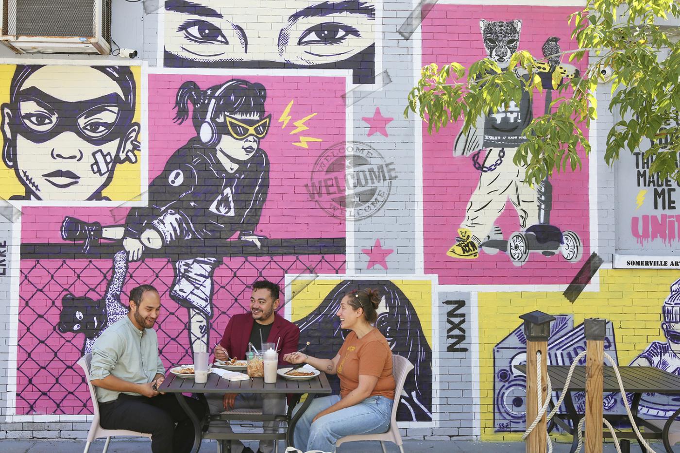 A mural of a collage of folks - a young girl climbs a fence, a woman wears a cat mask, a cat on a scooter, and eyes. Three folks sit at a table beneath it.