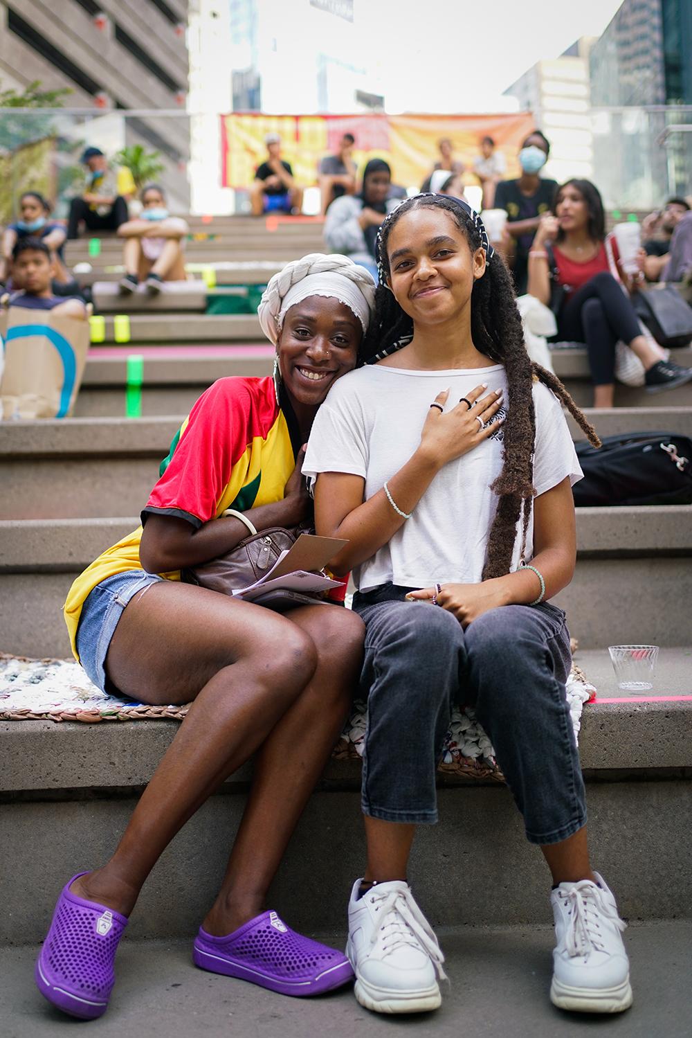 A young Black girl and an older Black woman pose together on steps. They snuggle and hold their hands to their chests.