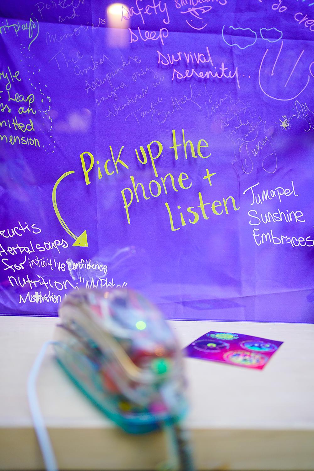 A multi-colored, chorded phone sits on a table in front of a sign that reads "pick up the phone and listen."